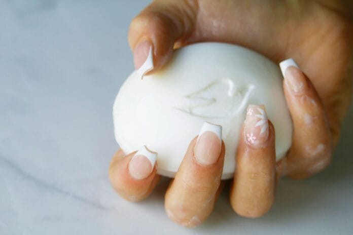 Dłoń z delikatnym french manicure trzymająca biały ozdobny kostkę mydła